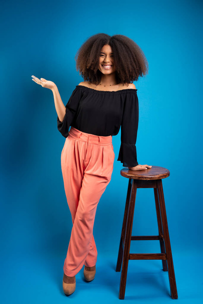 Młoda kobieta z czarnymi włosami, opierająca się na drewnianym stołku z dłonią skierowaną do góry. Izolacja na niebieskim tle. - Zdjęcie, obraz