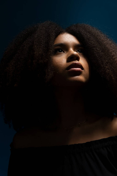 Half-closed portrait of young beautiful woman with black power hair looking up in low light. Isolated on blue background. - Photo, Image