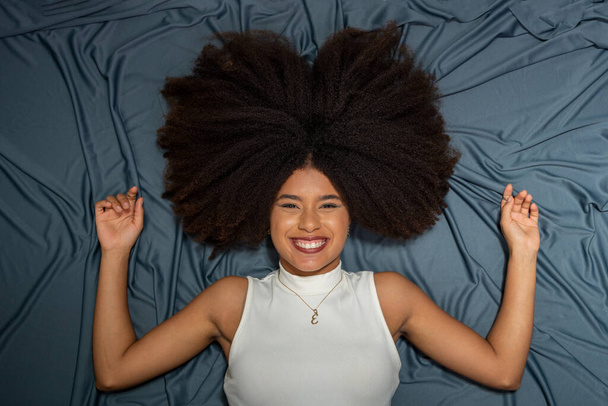 Beautiful young woman, with black power hair, smiling and looking at the camera lying on the floor with a gray background. - Photo, Image