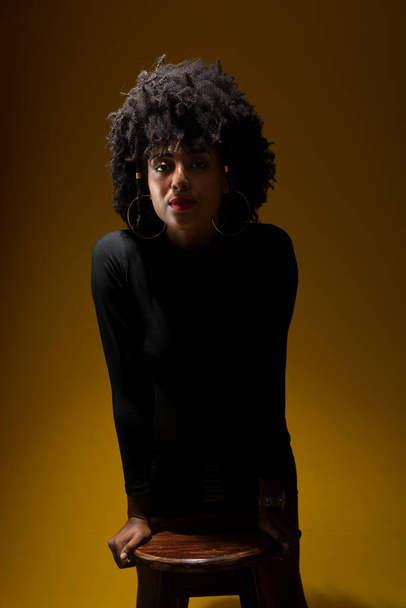 Portrait d'une jeune femme sensuelle aux cheveux noirs, portant des vêtements noirs posant pour une photo en basse lumière. Isolé sur fond jaune. - Photo, image