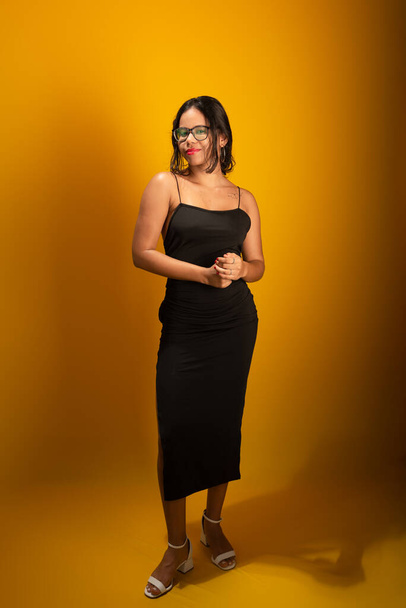 Porträt einer schönen sinnlichen jungen brünetten Frau, die schwarze Kleidung trägt, eine Brille trägt und für ein Foto posiert. Vereinzelt auf gelbem Hintergrund. - Foto, Bild