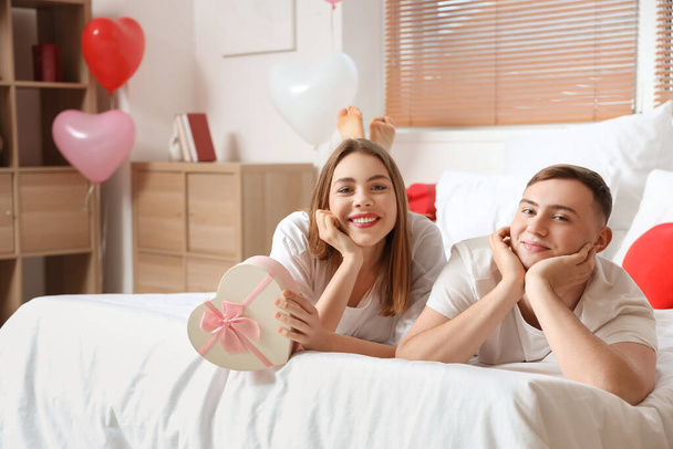 Beau jeune couple avec boîte cadeau couché sur le lit et célébrant la Saint-Valentin à la maison - Photo, image