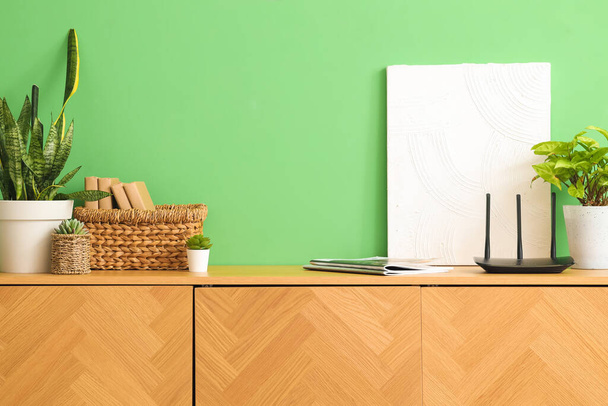 Roteador wi-fi moderno com plantas, pintura e livros em commode perto da parede verde - Foto, Imagem