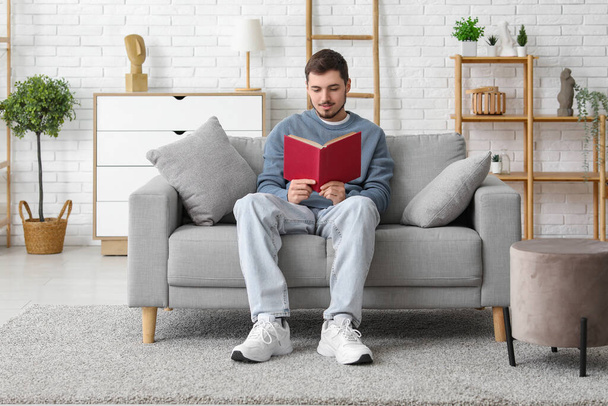 Jóképű fiatalember könyvet olvas, miközben a kanapén ül a nappaliban - Fotó, kép