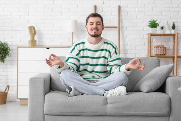 Schöner junger Mann meditiert auf Sofa im Wohnzimmer - Foto, Bild