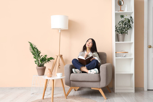 Ajattelevainen nuori aasialainen nainen, jolla on muistikirja, istuu nojatuolissa kotona - Valokuva, kuva