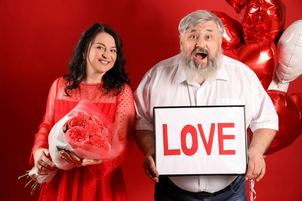 Krásný šokovaný dospělý pár s balónky ve tvaru srdce, kyticí růží a slovem LOVE na červeném pozadí. Valentýnská oslava - Fotografie, Obrázek