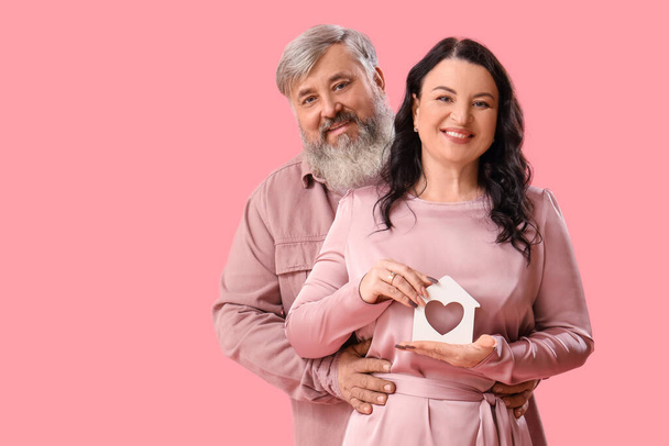 Beautiful mature couple holding figure of house with heart hugging on pink background. Valentine's Day celebration - Photo, Image