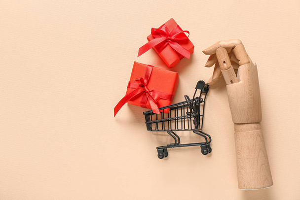 Small shopping cart with gift boxes and wooden hand on beige background. Valentine's Day celebration - Photo, Image