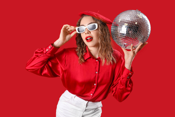 Mulher bonita em óculos de sol elegantes com bola de discoteca no fundo vermelho - Foto, Imagem