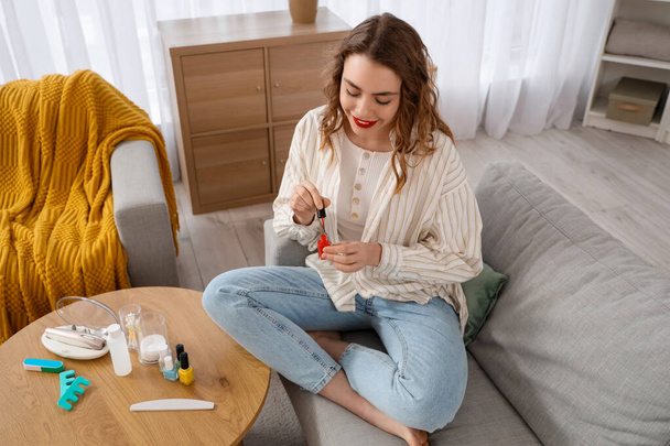 Schöne junge Frau mit Flaschen Nagellack und Zubehör für Maniküre sitzt im Wohnzimmer - Foto, Bild