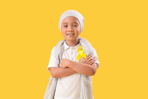 Cute little Asian girl after chemotherapy with yellow ribbon on color background. Childhood cancer awareness concept - Photo, Image