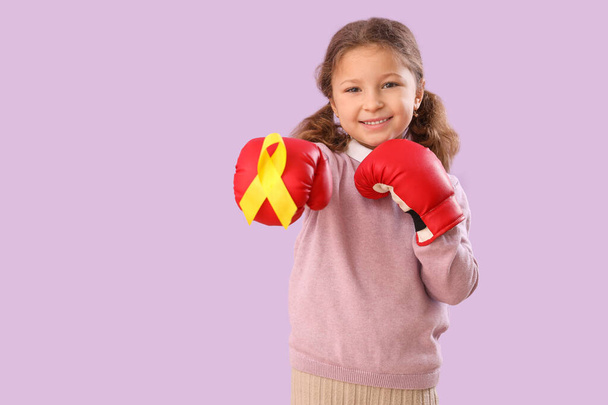 Cute little girl in boxing gloves with yellow ribbon on lilac background. Childhood cancer awareness concept - Photo, Image