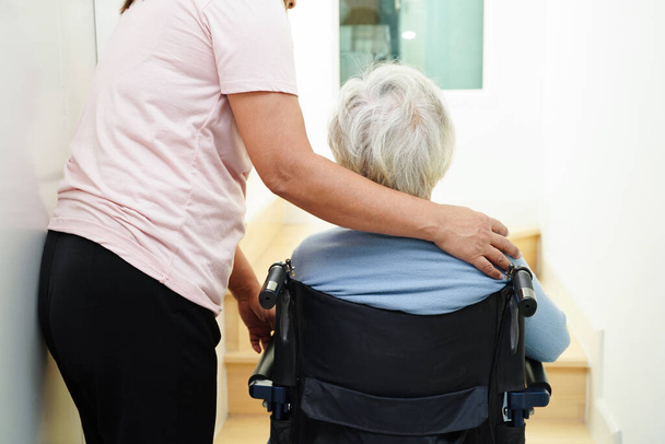 Mulher idosa asiática em cadeira de rodas com cuidador ajuda a apoiar a descer as escadas evitar acidentes, deslizamento e queda em casa. - Foto, Imagem