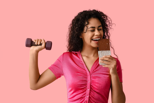 Красива молода афро-американка з солодким шоколадним баром і гантелі на рожевому фоні - Фото, зображення