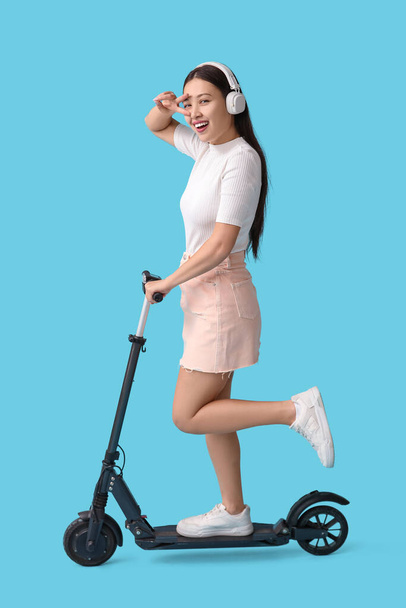 Happy young Asian woman in headphones riding modern electric kick scooter and showing victory gesture on blue background - Photo, Image