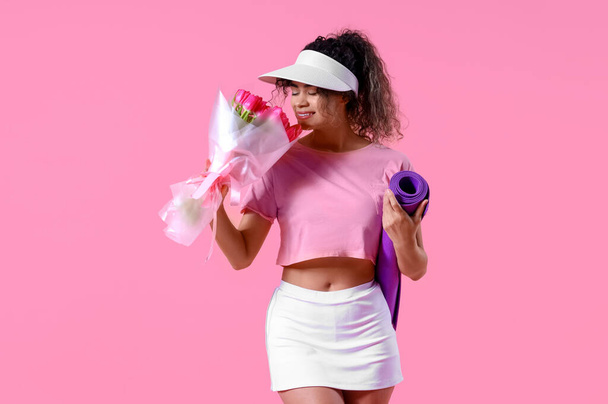 Young African-American woman in sportswear with bouquet of flowers and yoga mat on pink background. International Women's Day celebration - Photo, Image