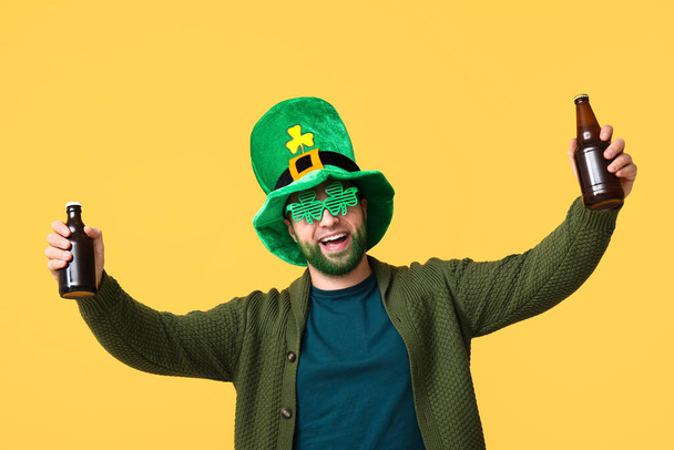 Szczęśliwy młody człowiek w karłowatym kapeluszu i ozdobnych okularach w kształcie koniczyny z zieloną brodą trzymający butelki piwa na żółtym tle. Święto św. Patryka - Zdjęcie, obraz