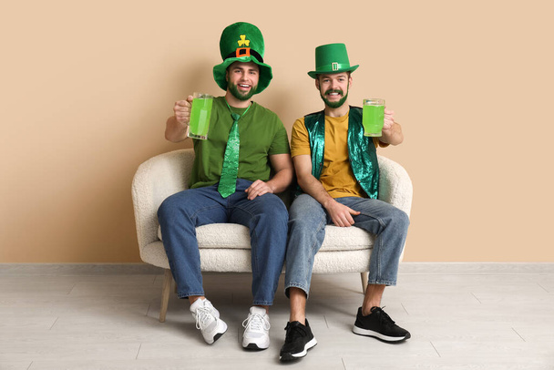 Giovani uomini felici in cappelli leprecauno con barbe verdi che tengono bicchieri di birra e seduti sul divano vicino alla parete beige. Festa di San Patrizio - Foto, immagini