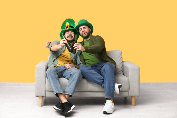 Молодые люди в лепреконовых шляпах с зелеными бородами держат бутылки пива и сидят на диване возле желтой стены. Празднование Дня Святого Патрика - Фото, изображение
