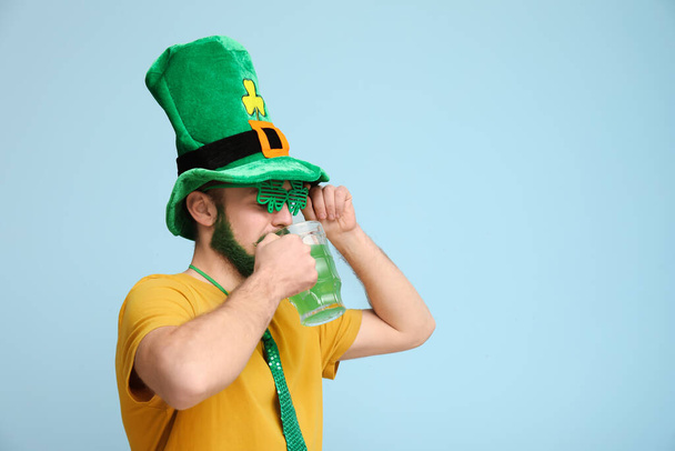 Leprikon şapkalı ve yeşil sakallı yonca şeklinde dekoratif gözlüklü genç adam mavi arka planda bira içiyor. Aziz Patrick Günü kutlaması - Fotoğraf, Görsel