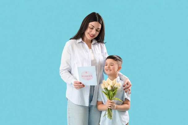Όμορφη γυναίκα και ο γιος της κρατώντας ευχετήρια κάρτα και μπουκέτο τουλίπες σε μπλε φόντο. Διεθνής Ημέρα της Γυναίκας - Φωτογραφία, εικόνα