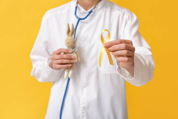 Doctor holding golden ribbon and toy bunny on yellow background. Childhood cancer awareness concept - Photo, Image