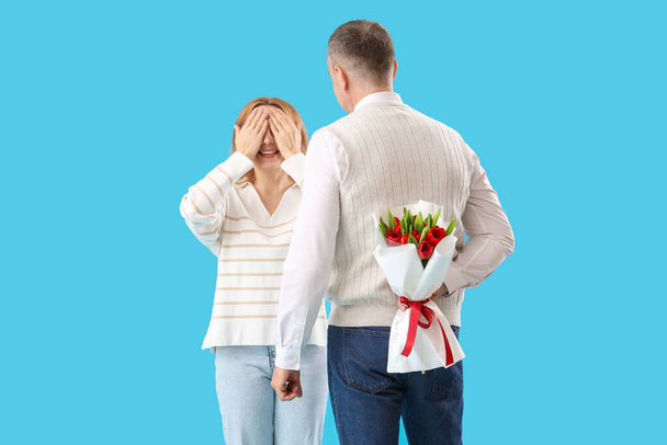 Hombre de mediana edad saludando a su esposa con ramo de flores sobre fondo azul. Día Internacional de la Mujer - Foto, Imagen