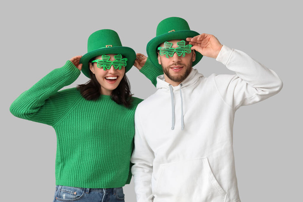 Красивая молодая пара в лепреконовых шляпах и декоративных очках в форме клевера на сером фоне. Празднование Дня Святого Патрика - Фото, изображение