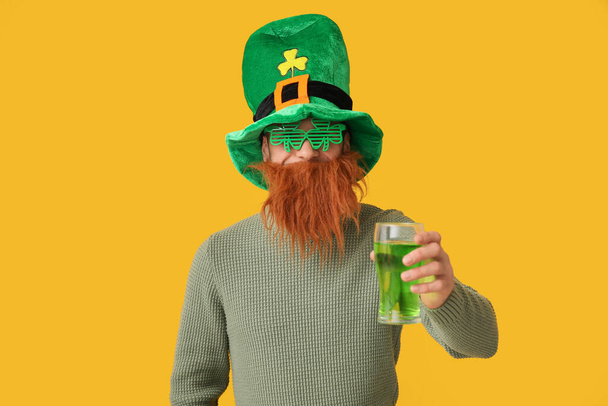 Смешной бородатый мужчина в шляпе лепрекона и декоративных очках в форме клевера со стаканом пива на желтом фоне. Празднование Дня Святого Патрика - Фото, изображение