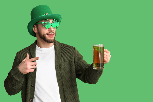 Egy kobold kalapos fiatalember, lóhere alakú díszítő szemüveggel, aki zöld hátterű sörre mutat. Szent Patrik napi ünnepség - Fotó, kép