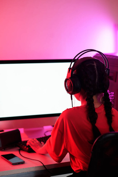Menina com duas tranças usando fone de ouvido ao jogar videogame no computador no quarto de néon, vista da parte de trás - Foto, Imagem