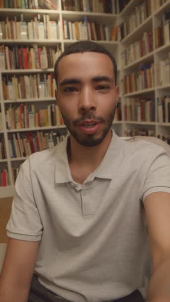 Medium vertical selfie shot of young Arab man sitting in public library among of bookcases with plenty of books, talking on smartphone camera and recording video for blog subscribers - Footage, Video