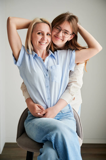 Słodka rodzinna chwila przytulania mamy i córki. Portret nastolatki pokazujący jej miłość do matki. Dwie kobiety w luźnych ciuchach przytulają się i patrzą w kamerę w domu. - Zdjęcie, obraz