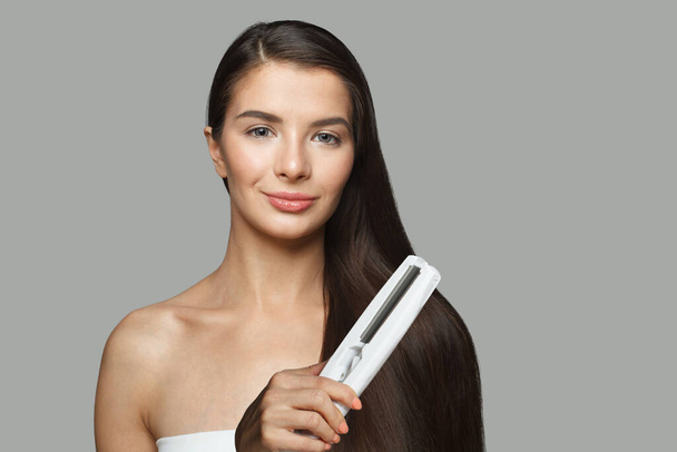 Μελαχρινή μοντέλο γυναίκα κρατώντας σίδερο για τα μαλλιά και ίσιωμα υγιή μακριά σκούρα μαλλιά της σε λευκό φόντο. Haircare και styling έννοια - Φωτογραφία, εικόνα