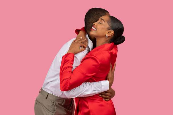 Чорний чоловік в хрусткій білій сорочці і жінка в яскраво-червоній сукні ділиться теплими, радісними обіймами, сміючись разом на рожевому фоні - Фото, зображення