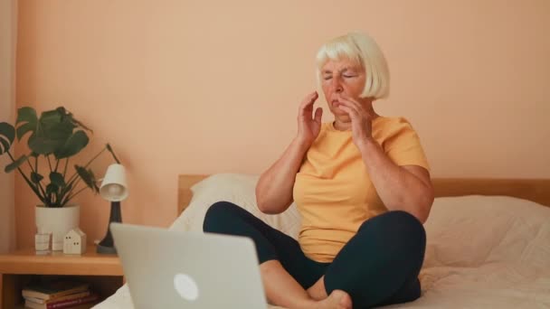 Retrato de la cansada mujer de 60 años con dolor de cabeza trabajó con el portátil en la cama en la habitación blanca en casa. Imágenes FullHD de alta calidad - Imágenes, Vídeo