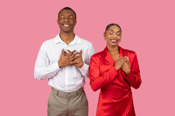 Schwarzes Paar in eleganter Kleidung mit Händen über Herzen und geschlossenen Augen, Dankbarkeit und Zufriedenheit ausdrückend, posierend auf rosa Hintergrund - Foto, Bild