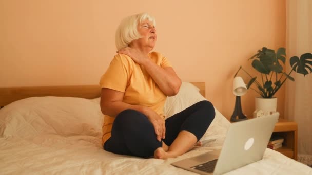 Retrato de la cansada mujer de 60 años con dolor de cabeza trabajó con el portátil en la cama en la habitación blanca en casa. Mujer de 50 años con dolor de hombro, sentada en pijama en su cama. La anciana está soportando - Imágenes, Vídeo
