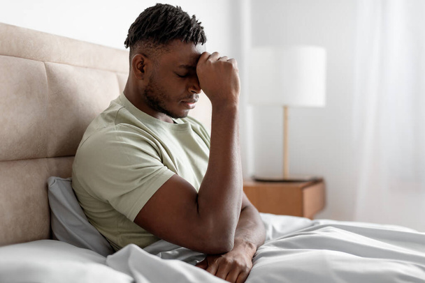 Unhappy black young man in bed pressing fist to forehead suffering from depression and chronic headache problem, waking up in the morning in modern bedroom at home, side view - Photo, Image