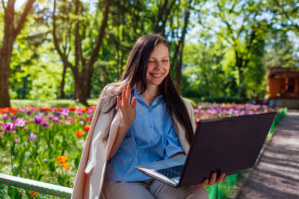 Außenporträt einer glücklichen Geschäftsfrau, die im Frühlingspark am Laptop arbeitet. Frau telefoniert per Videokonferenz und winkt Cowokern im öffentlichen Garten zu - Foto, Bild
