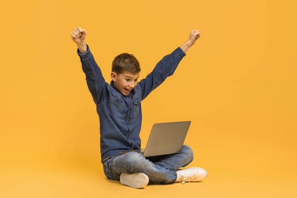 Opgewonden jongen met laptop vieren succes tijdens het zitten op de vloer in de studio, vrolijke tiener mannelijke kind het verhogen van de armen in de overwinning pose, het overbrengen van prestatie of goed nieuws, geïsoleerd op gele achtergrond - Foto, afbeelding