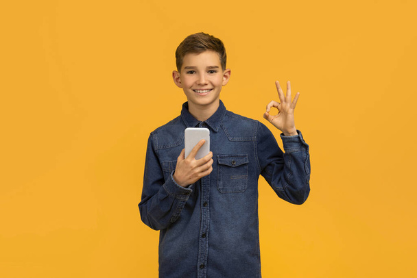 Ευτυχισμένος έφηβος αγόρι κρατώντας smartphone και κάνοντας OK χειρονομία με το χέρι, χαμογελώντας έφηβος αρσενικό παιδί κρατώντας κινητό τηλέφωνο, συνιστώντας νέα εφαρμογή για κινητά, στέκεται κατά κίτρινο φόντο στούντιο, αντίγραφο χώρο - Φωτογραφία, εικόνα