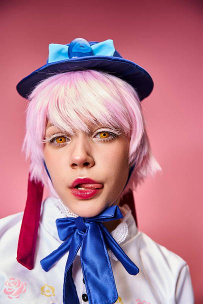verführerische junge Cosplayerin streckt ihre Zunge heraus und blickt vor rosa Hintergrund in die Kamera - Foto, Bild