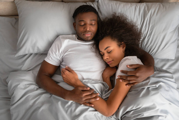 Nagy látószögű kilátás szeretetteljes afro-amerikai házastársakra, akik ölelkeznek és ölelgetik egymást, miközben kényelmes ágyban alszanak. Az éjszakai pihenés alatt ölelkező szerető fiatal család feje fölött - Fotó, kép