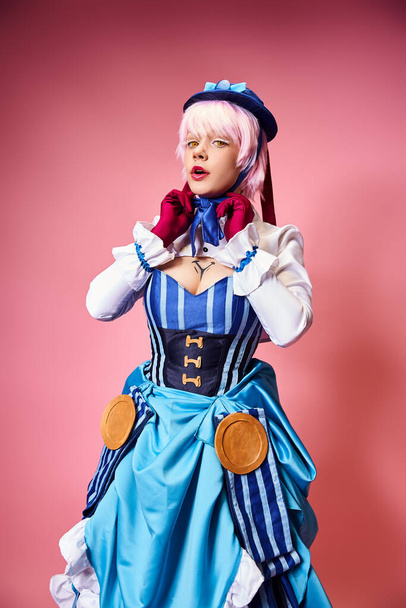 enticing sexy female cosplayer in blue hat and vibrant attire looking at camera on pink backdrop - Zdjęcie, obraz