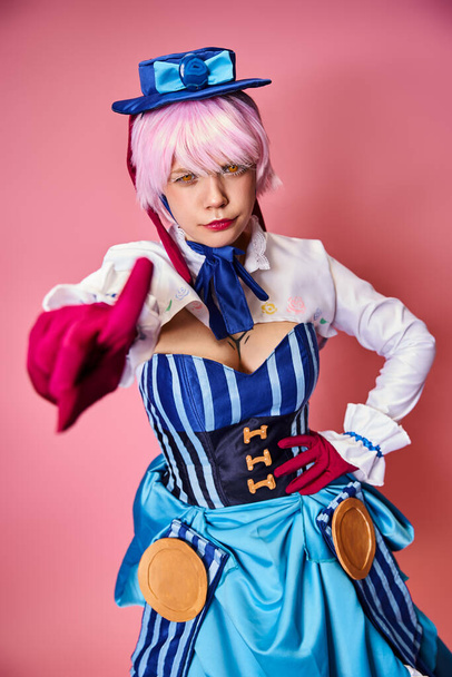 attractive female cosplayer in vibrant dress with blue hat pointing at camera on pink backdrop - Φωτογραφία, εικόνα