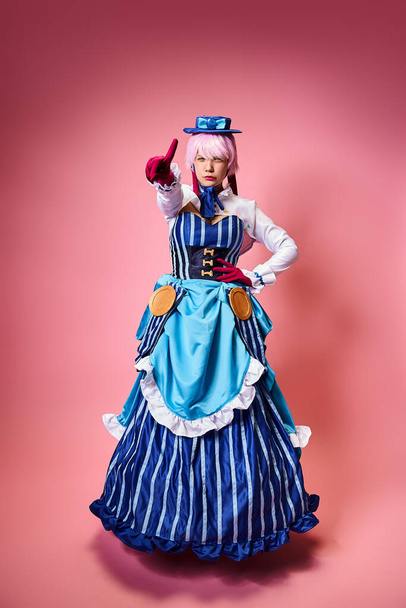 alluring female cosplayer in vibrant dress with blue hat pointing at camera on pink backdrop - Foto, Bild
