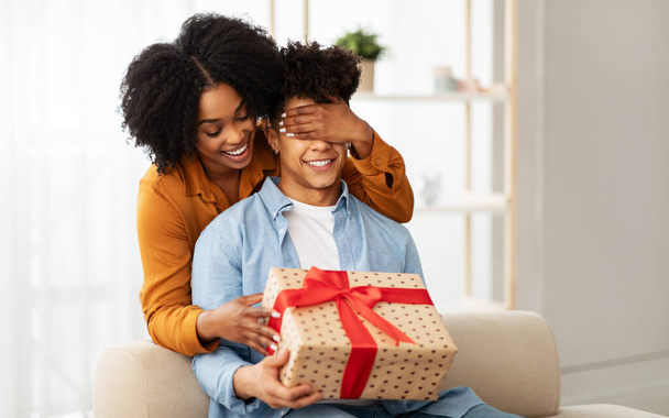 Eine fröhliche afrikanisch-amerikanische Frau verdeckt spielerisch die Augen eines Mannes von hinten und beide lächeln voller Vorfreude, während sie eine große, herzgemusterte Geschenkbox mit einer roten Schleife in der Hand halten. - Foto, Bild