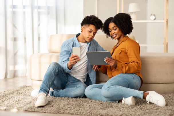 Casualmente vestido de ganga, um jovem casal afro-americano senta-se em um tapete de pelúcia, absorvido em seus dispositivos, com o homem segurando um telefone e a mulher um tablet em um ambiente sereno em casa - Foto, Imagem